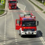 Red fire engine with blue emergency lights drives down a two-lane road, side signs and a pedestrian crossing sign visible; gear on top, open driver's door, officer nearby.