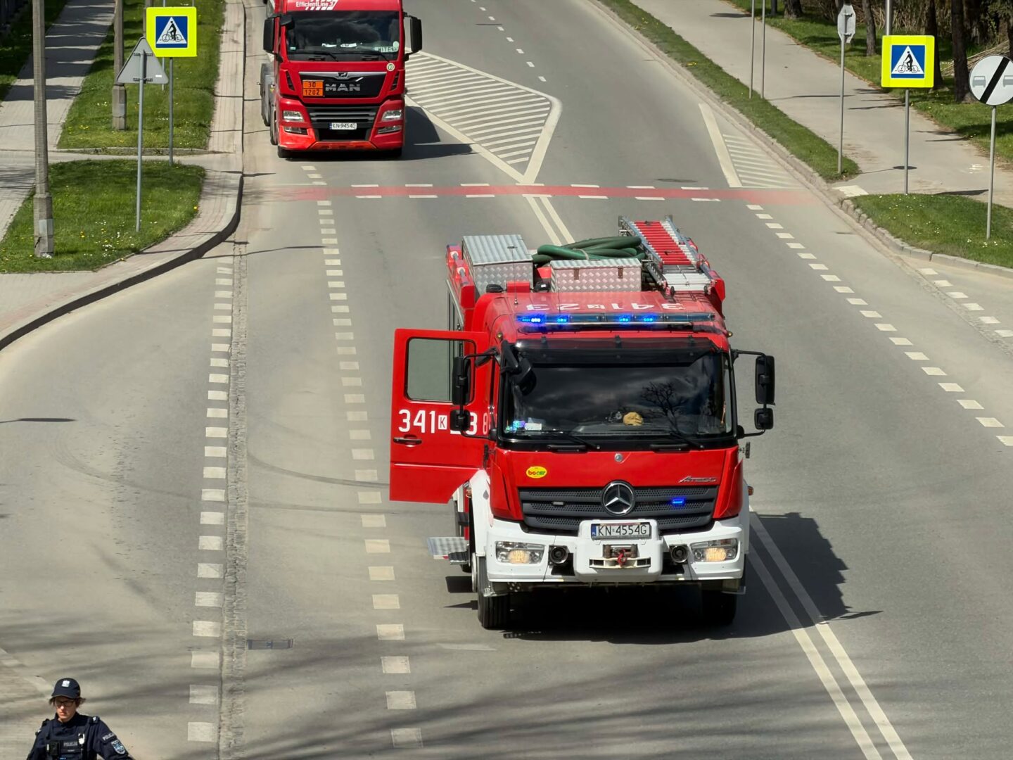 Red fire engine with blue emergency lights drives down a two-lane road, side signs and a pedestrian crossing sign visible; gear on top, open driver's door, officer nearby.