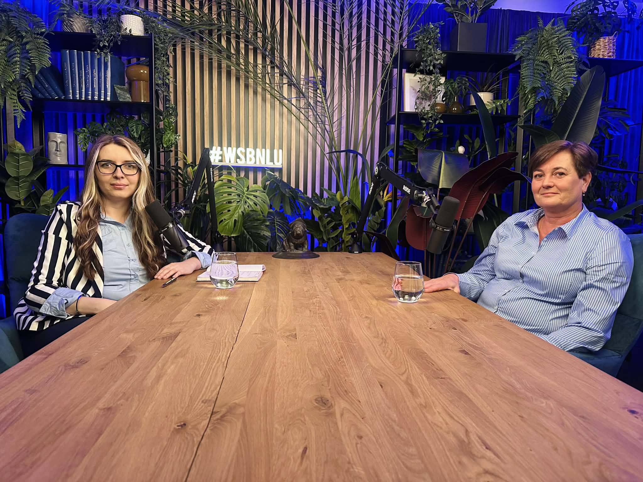 Two women sit at a wooden table with microphones in a plant-filled studio, blue lighting, and a hashtag sign in the background.