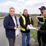 Four men on a roadside discussing documents; two wear high-visibility vests, one holds a clipboard and notes under a 30 km/h sign.
