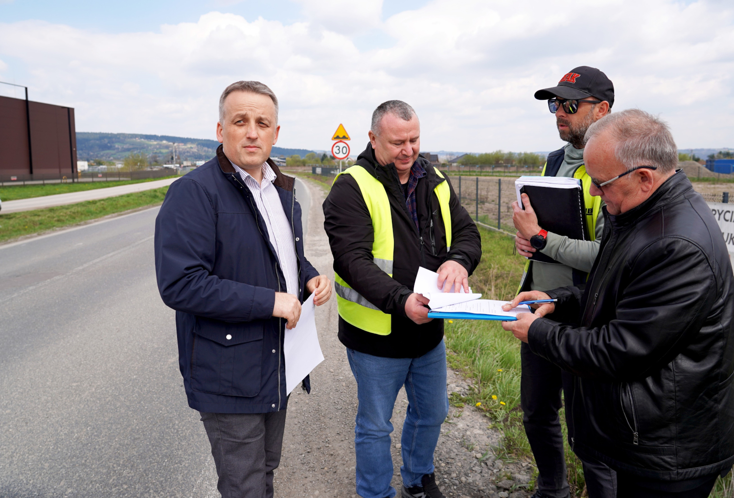 Four men on a roadside discussing documents; two wear high-visibility vests, one holds a clipboard and notes under a 30 km/h sign.