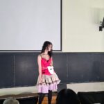 Young woman in a pink corset top and layered skirt stands on a classroom theater stage; a white mask hangs from her waist.