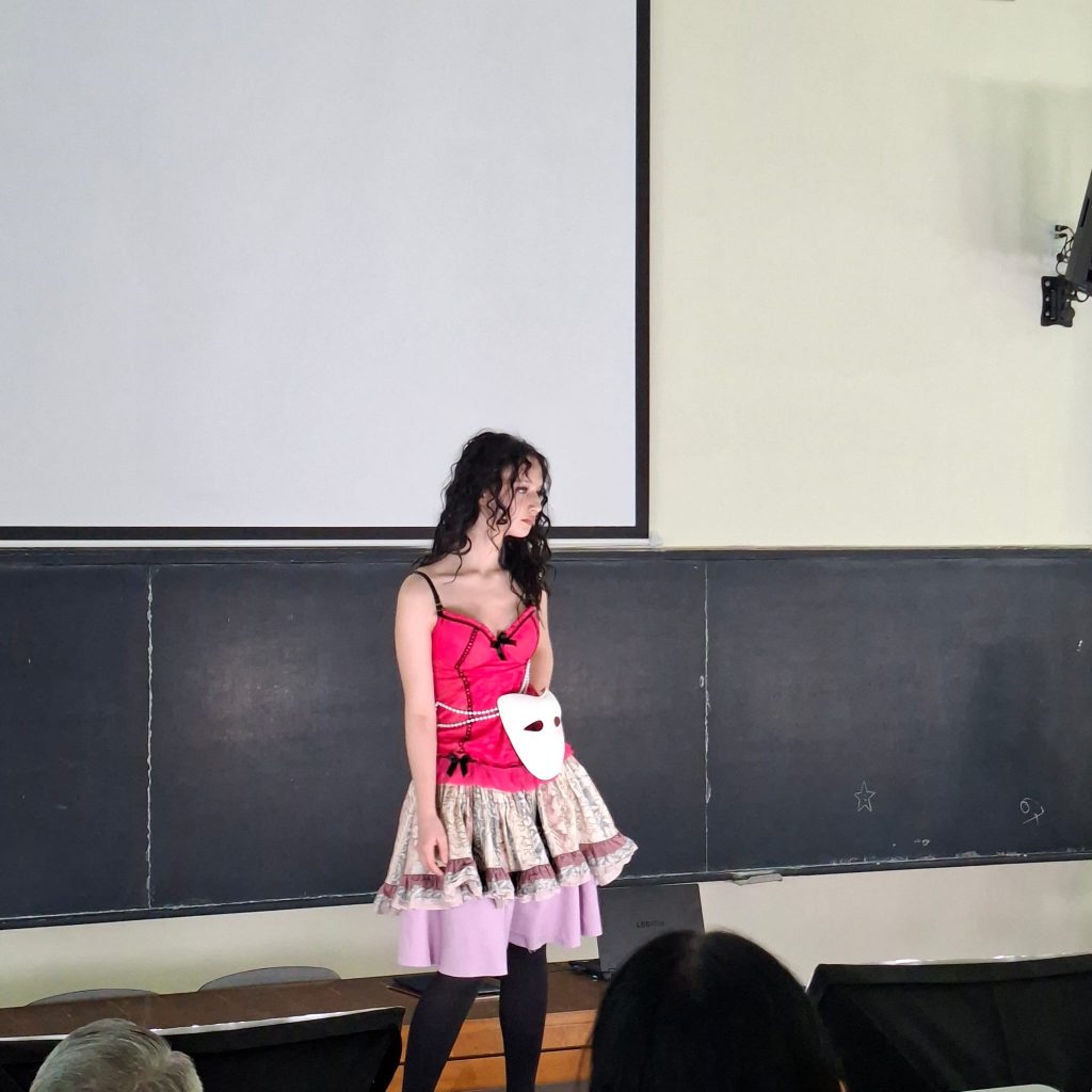 Young woman in a pink corset top and layered skirt stands on a classroom theater stage; a white mask hangs from her waist.