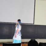 Girl in a blue and white costume with braided hair stands at a podium or desk in a classroom, holding a flower, facing left in front of a large screen and chalkboard railings.