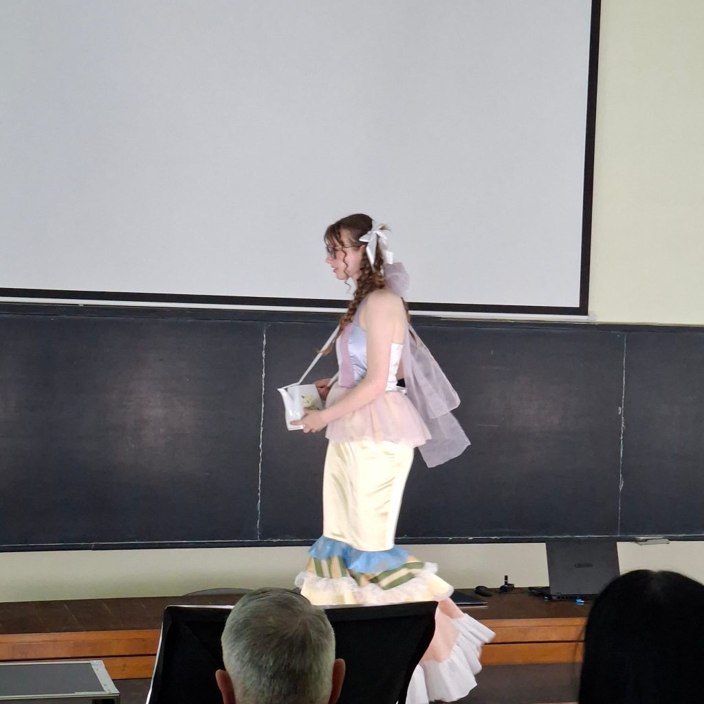 Person in pastel ballerina costume walking on a stage, holding a white bucket, chalkboard behind, audience seating in foreground