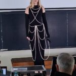 Female model in a black cold-shoulder outfit with white rope-like accessories stands on a stage in front of a chalkboard, audience seated in a classroom.