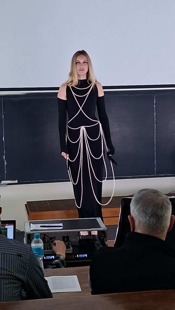 Female model in a black cold-shoulder outfit with white rope-like accessories stands on a stage in front of a chalkboard, audience seated in a classroom.