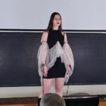 Model on a stage wearing a black mini dress with a sheer gray cape, in front of a chalkboard; audience member visible in foreground.