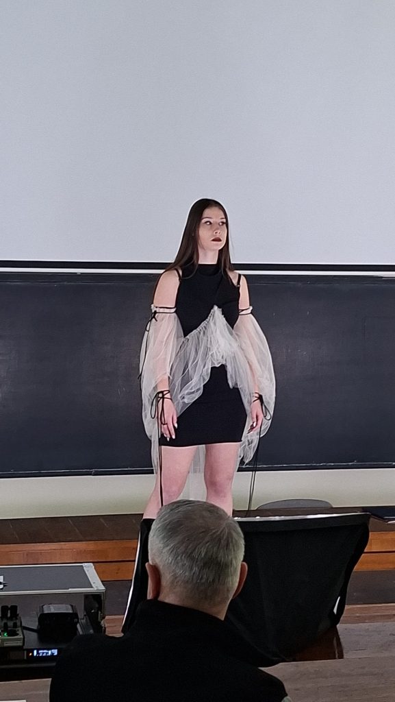 Model on a stage wearing a black mini dress with a sheer gray cape, in front of a chalkboard; audience member visible in foreground.