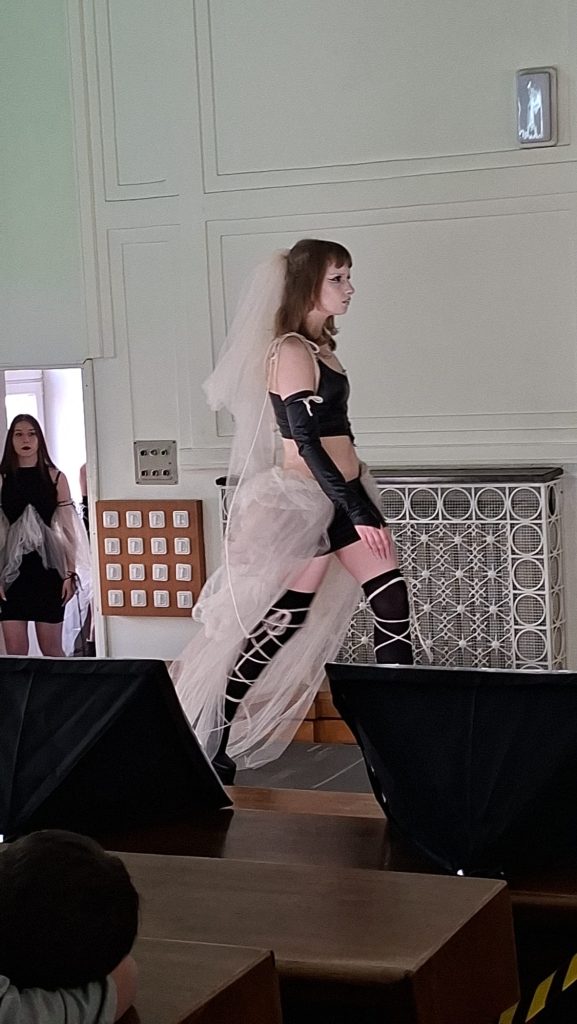 Model walking a runway in a black two-piece outfit with a white veil and long gloves, in a formal venue with audience seating nearby.