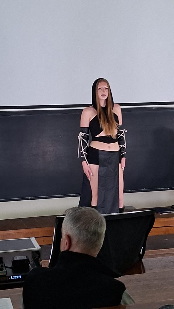 Young woman in a black, cut‑out outfit with lace-up sleeves standing on a stage in front of a chalkboard, audience visible in the foreground