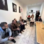 People sit in a meeting room along a white wall with portraits; a speaker stands at a long wooden table to the right.