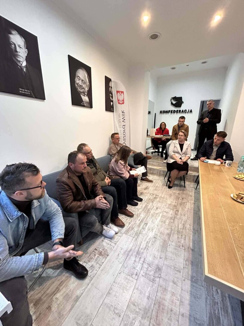 People sit in a meeting room along a white wall with portraits; a speaker stands at a long wooden table to the right.