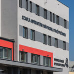 Facade of a modern Polish fire service training building with bold blue letters: 'SZKOŁA ASPIRANTÓW PAŃSTWOWEJ STRAŻY POŻARNEJ' and 'Wydział Szkoleń Specjalistycznych' against a clear blue sky, with red-framed windows on the lower level.