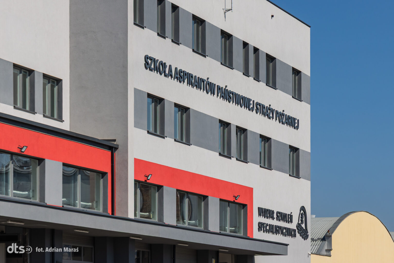Facade of a modern Polish fire service training building with bold blue letters: 'SZKOŁA ASPIRANTÓW PAŃSTWOWEJ STRAŻY POŻARNEJ' and 'Wydział Szkoleń Specjalistycznych' against a clear blue sky, with red-framed windows on the lower level.