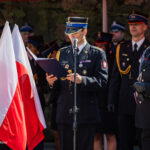 Polish military officers in dress uniforms at a flag ceremony, one reading from a folder at a microphone with a white-and-red flag in the foreground.