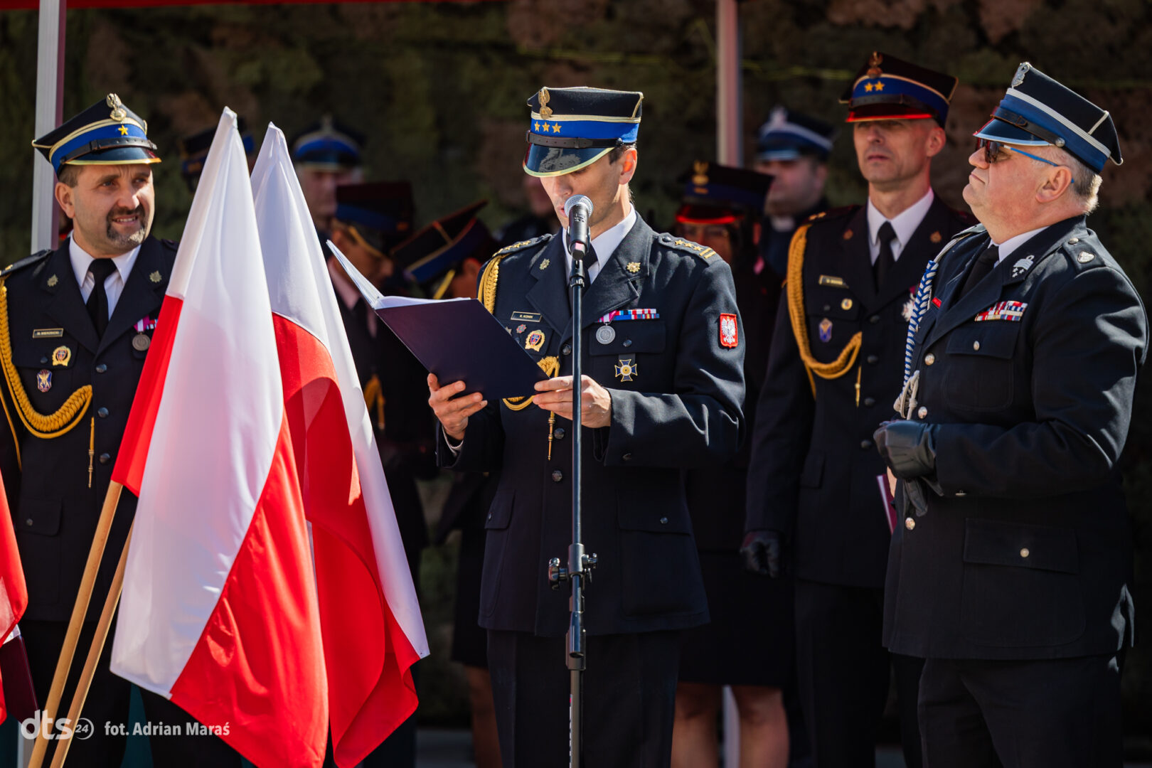 Polish military officers in dress uniforms at a flag ceremony, one reading from a folder at a microphone with a white-and-red flag in the foreground.