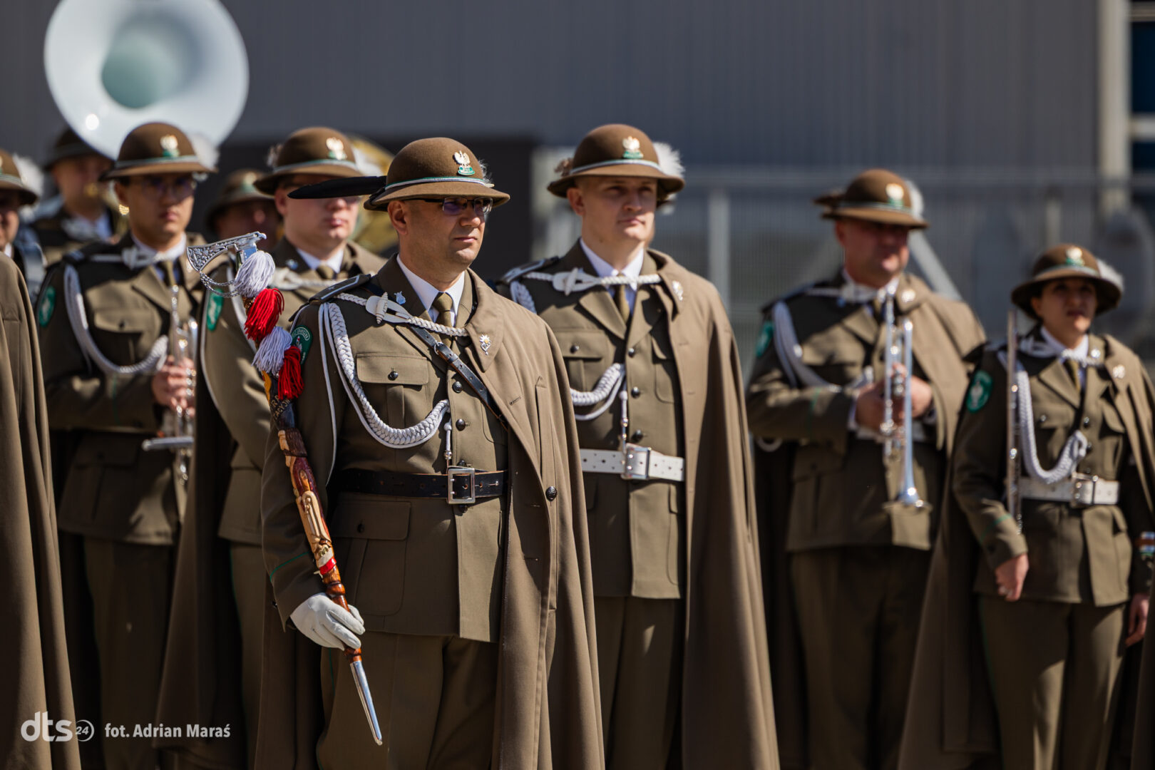Ceremonial soldiers in brown uniforms with plumed hats, standing with swords and braided cords during a formal parade
