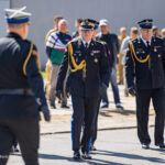 Military officers in dress uniform walking on a tarmac, gold cords and medals visible, formation of personnel in background (ceremonial event).