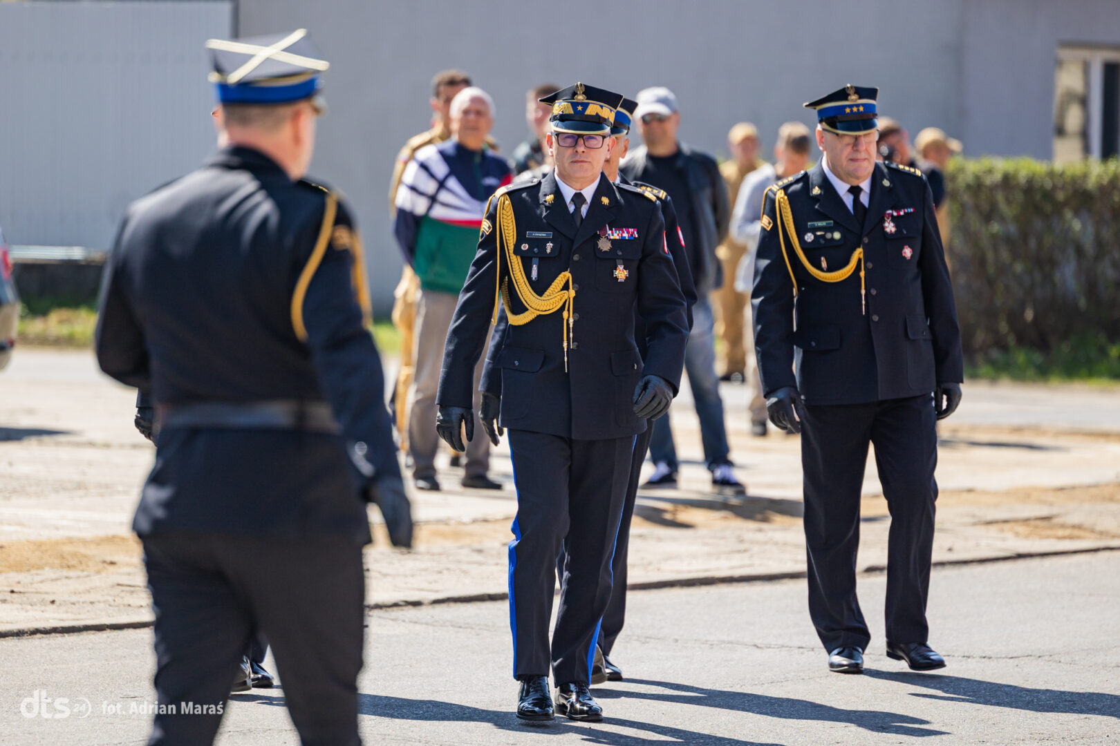 Military officers in dress uniform walking on a tarmac, gold cords and medals visible, formation of personnel in background (ceremonial event).
