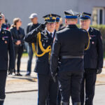 Four military officers in dress blues with gold cords stand on a road; central officer salutes a colleague facing him, while others look on.