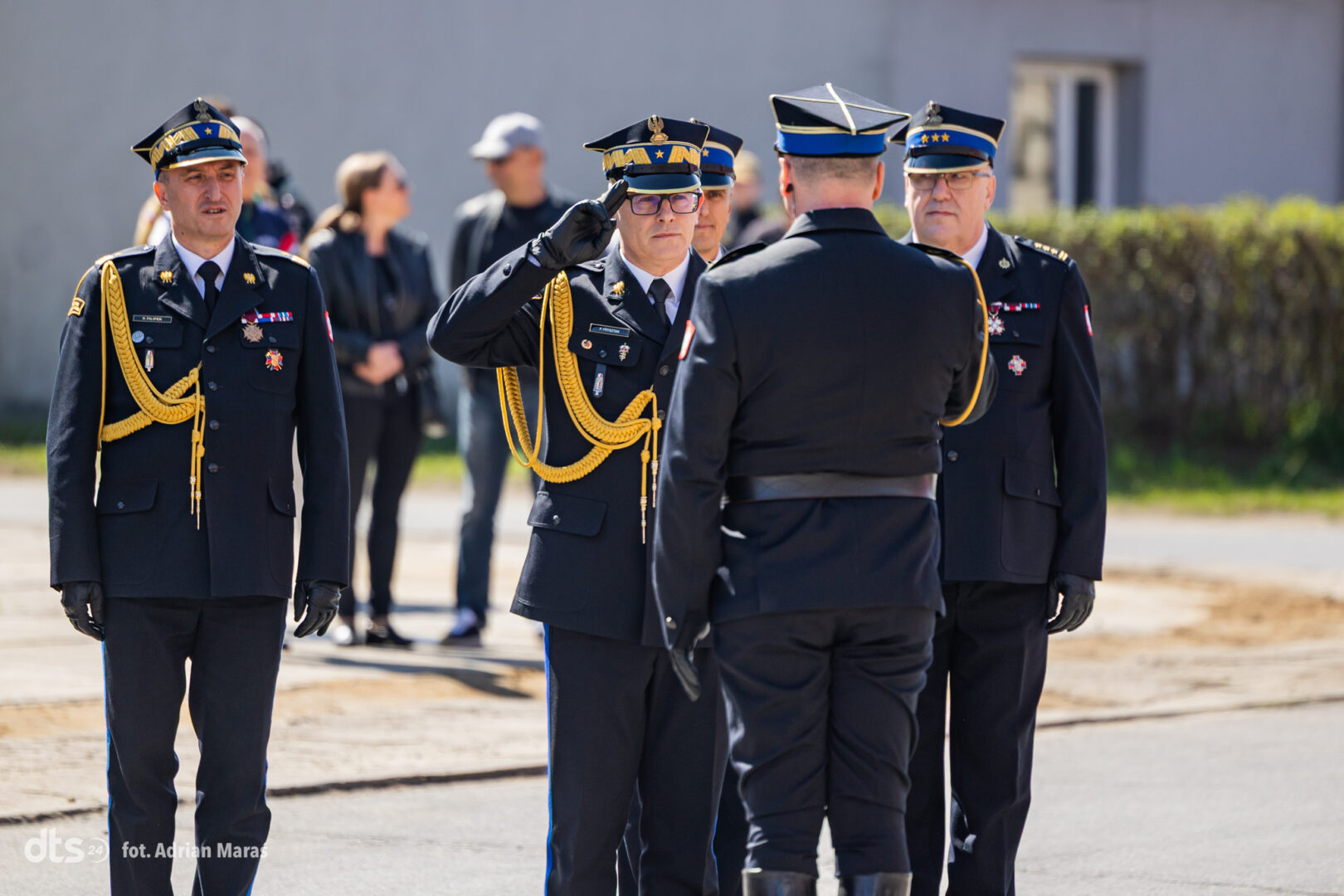 Four military officers in dress blues with gold cords stand on a road; central officer salutes a colleague facing him, while others look on.