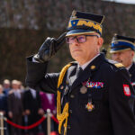 Polish army officer in dress uniform saluting at an outdoor ceremony, medals on chest with a crowd in the background.