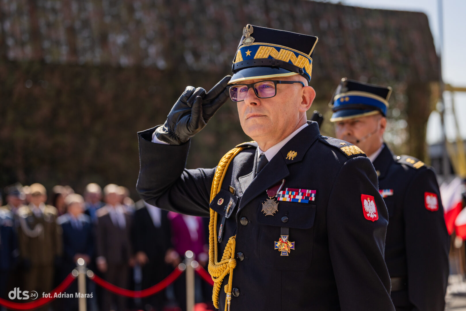 Polish army officer in dress uniform saluting at an outdoor ceremony, medals on chest with a crowd in the background.