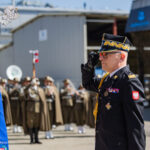 Polish military officer in dress uniform salutes beside a ceremonial flag at a parade.