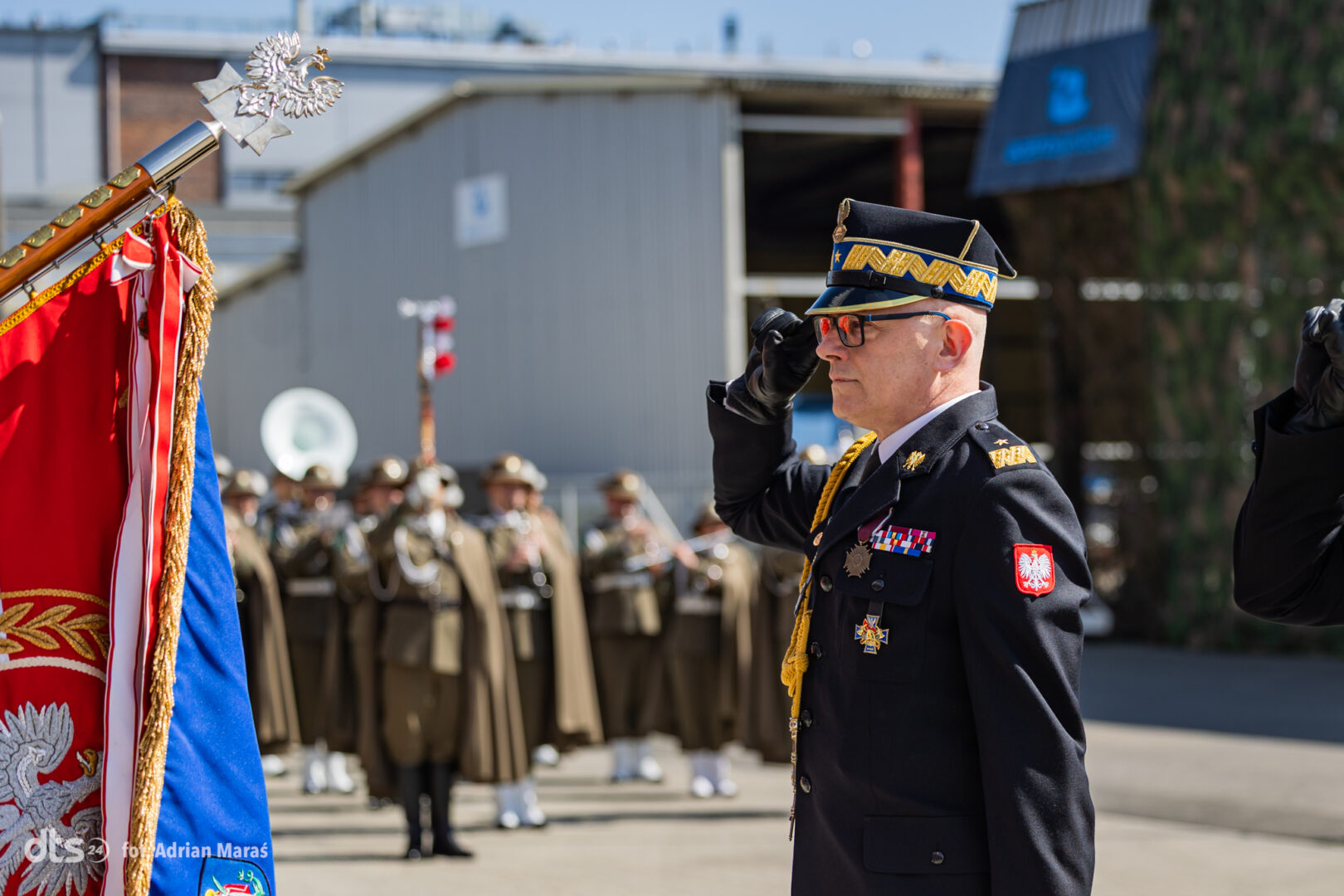 Polish military officer in dress uniform salutes beside a ceremonial flag at a parade.