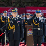 Senior military officer in ceremonial uniform speaks at a microphone, while officers stand in formation behind him on a red carpet.