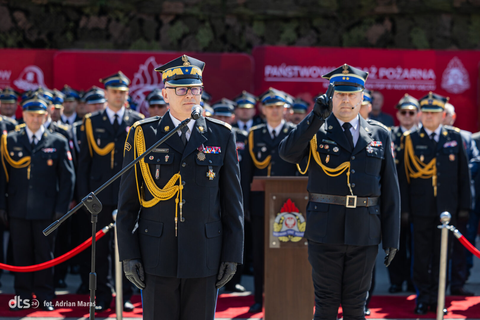Senior military officer in ceremonial uniform speaks at a microphone, while officers stand in formation behind him on a red carpet.