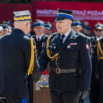 Two high-ranking military officers in dress uniforms shake hands during a formal ceremony, with medals and gold cords, as other officers stand in formation behind them.
