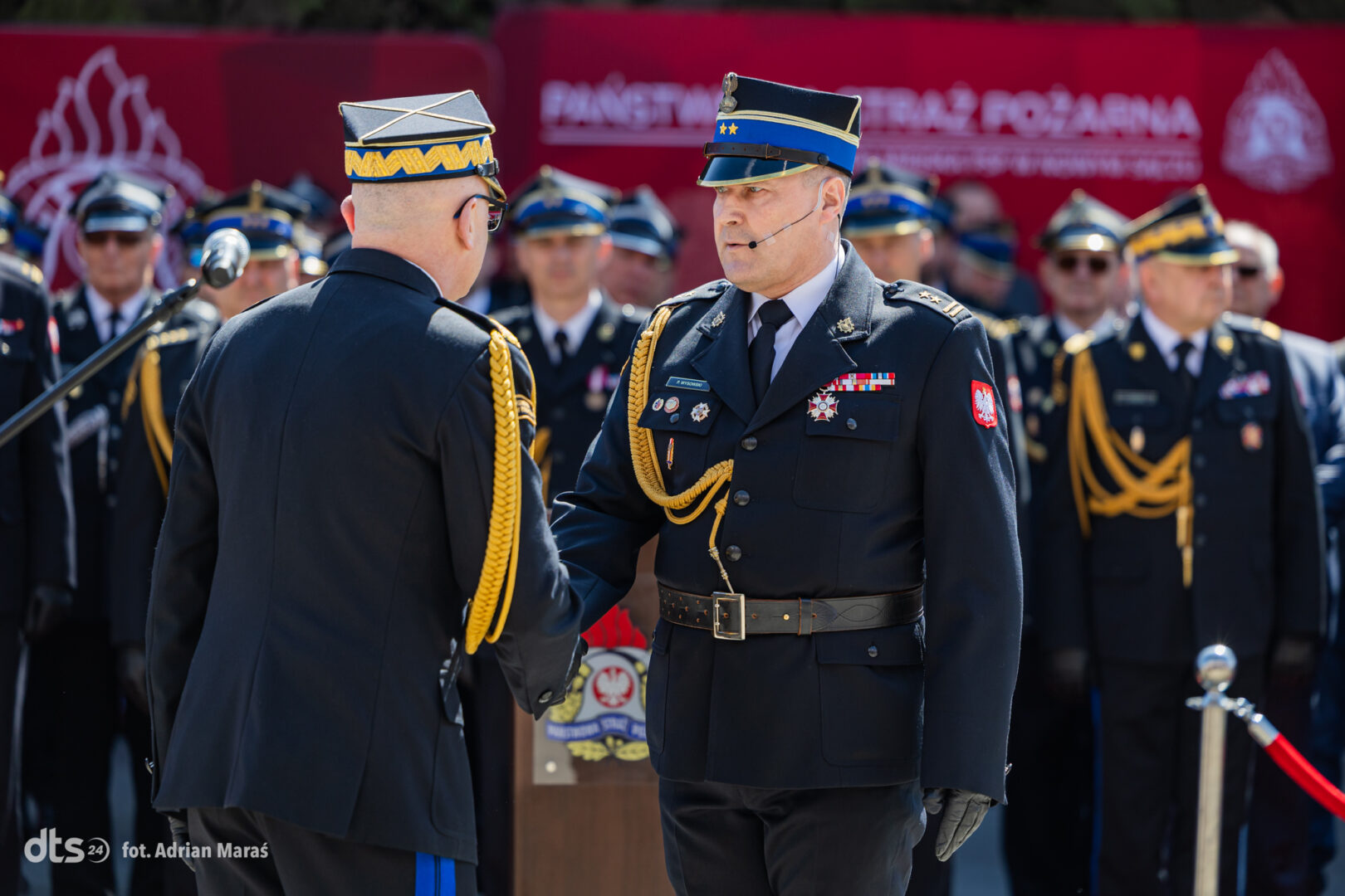 Two high-ranking military officers in dress uniforms shake hands during a formal ceremony, with medals and gold cords, as other officers stand in formation behind them.