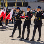 Three Polish military cadets in formal dress uniforms march in formation, carrying a folded red-and-white flag, with onlookers and flags in the background.