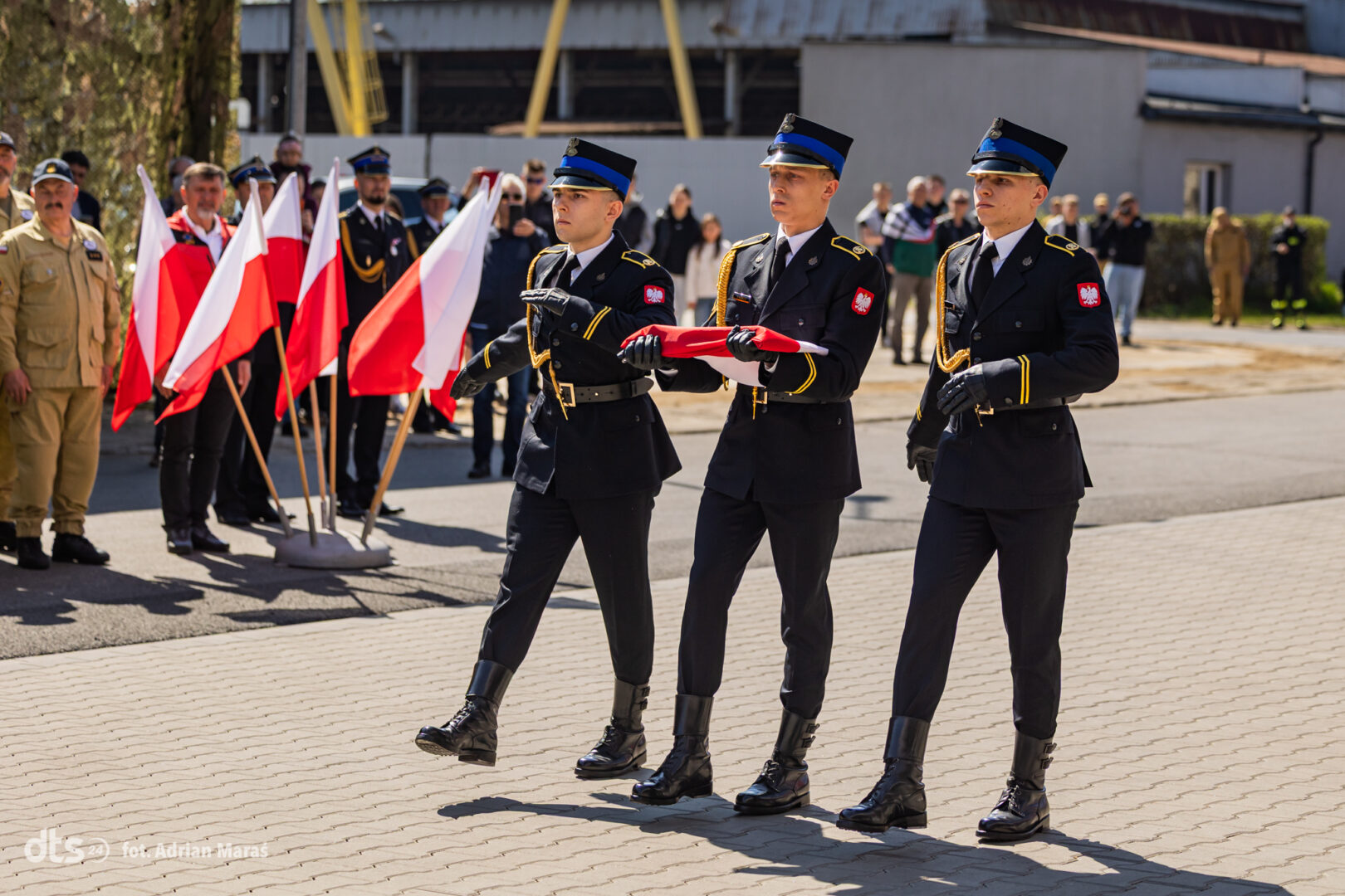 Three Polish military cadets in formal dress uniforms march in formation, carrying a folded red-and-white flag, with onlookers and flags in the background.