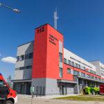 Red fire station tower labeled 112 with a gray and white building facade on a sunny day; ambulances and fire trucks parked outside.