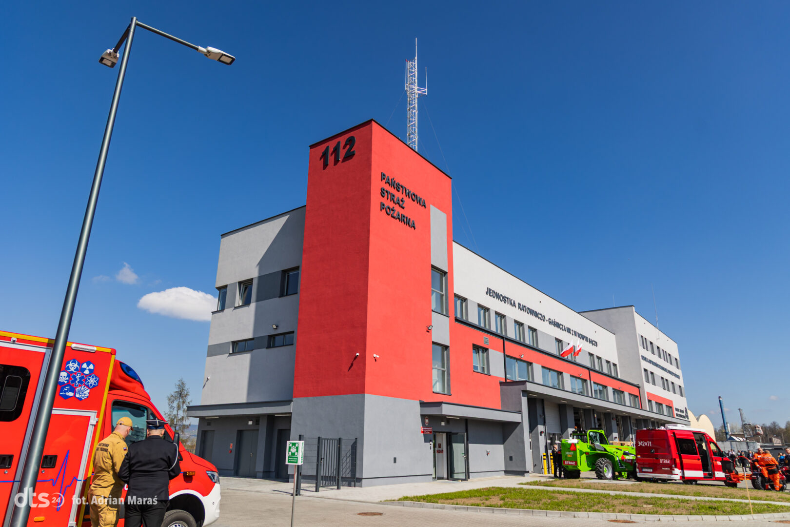 Red fire station tower labeled 112 with a gray and white building facade on a sunny day; ambulances and fire trucks parked outside.