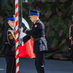 Three uniformed soldiers participate in a flag‑raising ceremony, one handling the flagpole while another assists and a third stands at attention in the background.