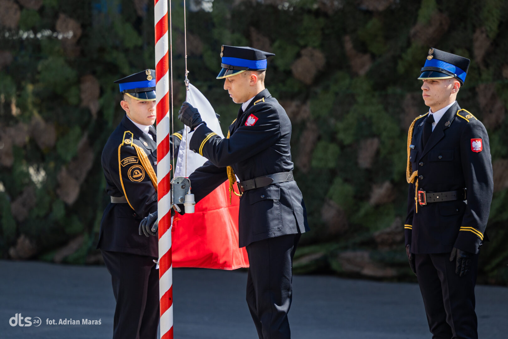 Three uniformed soldiers participate in a flag‑raising ceremony, one handling the flagpole while another assists and a third stands at attention in the background.