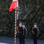 Three guards in formal uniforms stand at attention as one salutes near a tall red-and-white striped flagpole during a ceremony.
