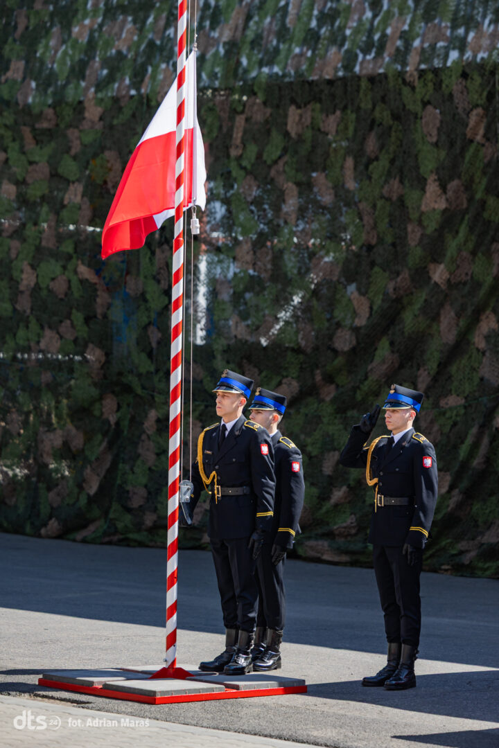 Three guards in formal uniforms stand at attention as one salutes near a tall red-and-white striped flagpole during a ceremony.