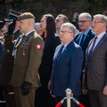 Polish military officer saluting in olive uniform during a formal ceremony, with officials in suits and other uniformed personnel nearby.