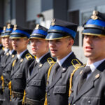 Line of military cadets in black dress uniforms with blue peaked caps and gold trim, standing at attention.