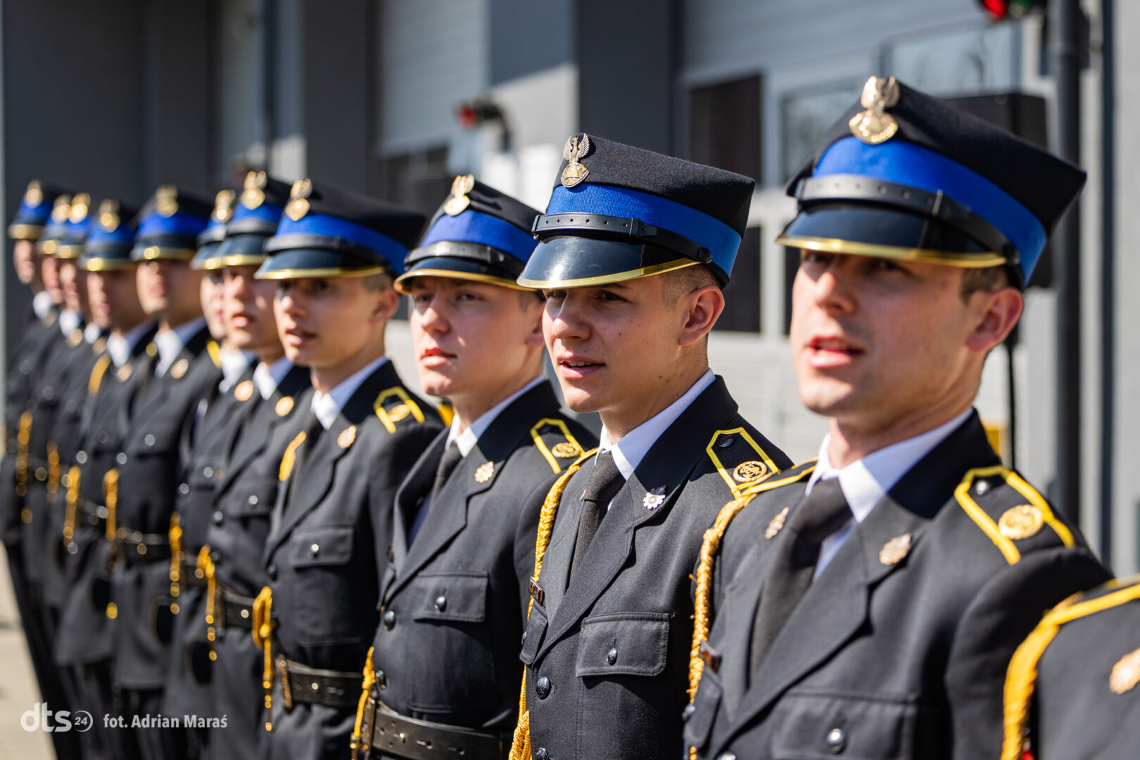 Line of military cadets in black dress uniforms with blue peaked caps and gold trim, standing at attention.