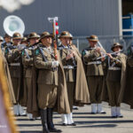 Military band in brown uniforms marching outdoors, playing brass and woodwind instruments in formation.