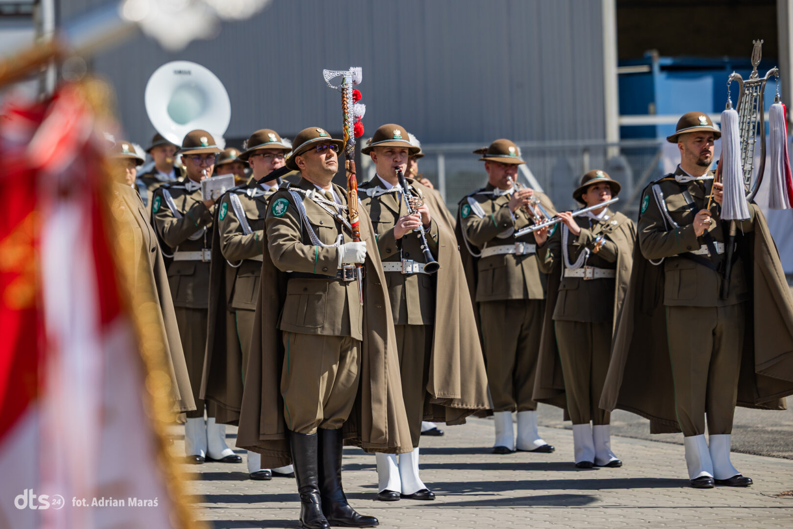 Military band in brown uniforms marching outdoors, playing brass and woodwind instruments in formation.