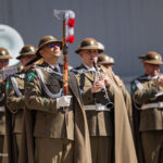 Military band in olive uniforms outdoors; lead officer holds a decorated ceremonial baton with red tassels.