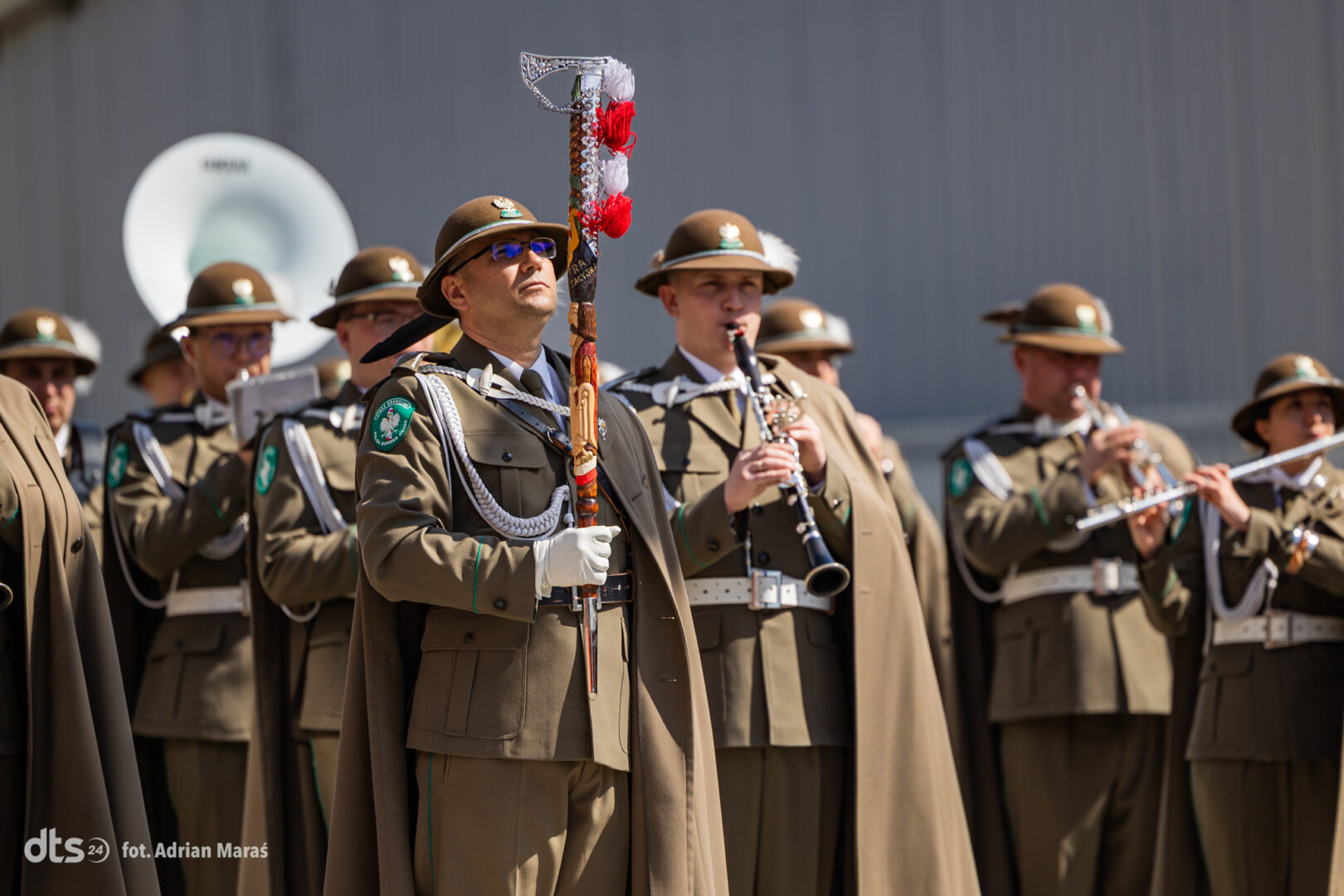 Military band in olive uniforms outdoors; lead officer holds a decorated ceremonial baton with red tassels.