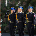 Three uniformed soldiers salute beside a striped pole during a ceremony, Poland patch visible on one sleeve.